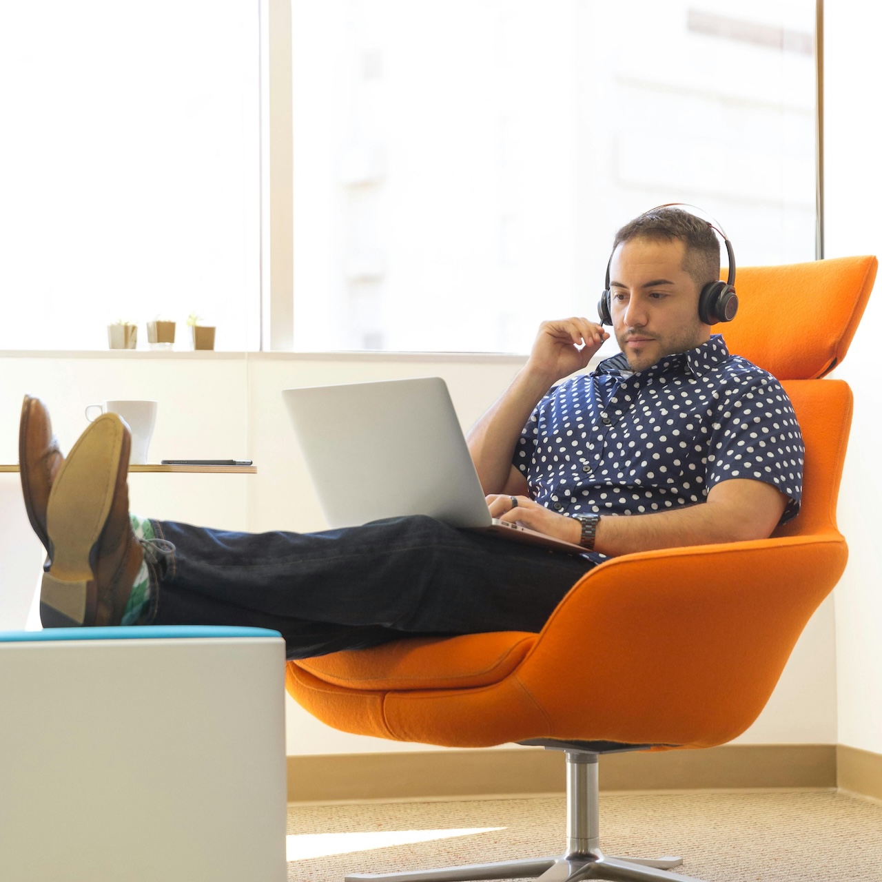 Man with laptop and headphones lounging on an orange chair with his feet on a footstool